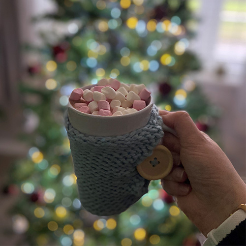 A grey mug topped with mini marshmallows held up in front of a Christmas tree