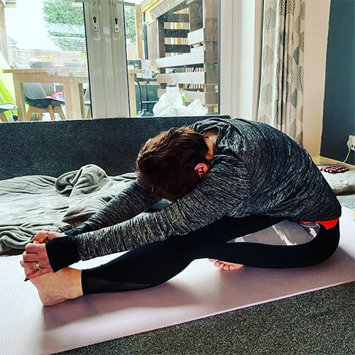 Slimming World member Kate wearing grey gym clothes, sitting on a pink yoga mat, stretching to touch her toes