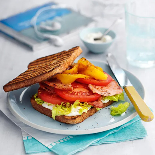 Slimming World Bacon, lettuce and tomato sandwich. The sandwich is made up of two slices of toasted wholemeal bread filled with lettuce, slices of tomato, bacon and yellow peppers. It sits on a light blue plate, which rests on a white tabletop.