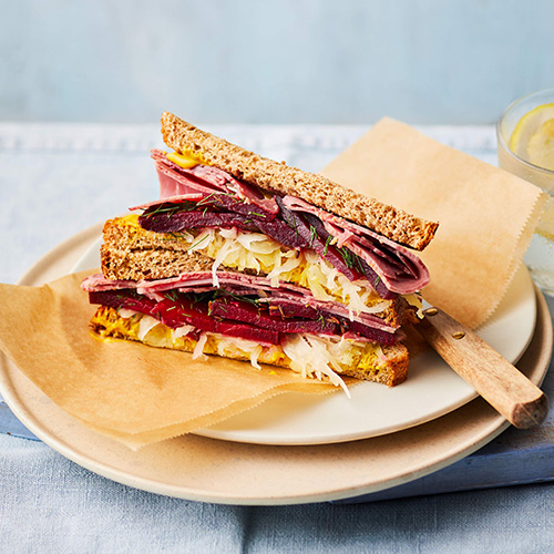Slimming World New York deli-style salt beef and mustard sandwich. The sandwich is made up of two slices of wholemeal bread filled with beef, sauerkraut and marinated beetroot. It sits on light brown baking parchment on top of a white plate. The background is light grey.